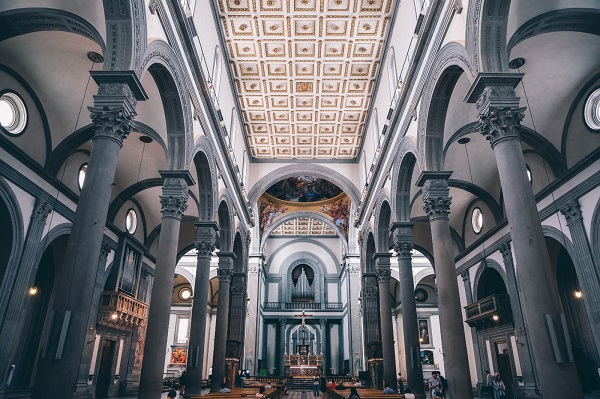 La foto riprende l’interno della basilica di San Lorenzo a Firenze come se la vedessimo stando nella navata centrale. Al lato destro e sinistro due file di alte colonne segnano le navate laterali. Gli archi tra le colonne compongono i moduli quadrati delle campate che definiscono lo spazio in modo ritmico e equilibrato