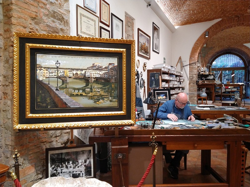 nell'immagine si vede la bottega e negozio di scarpelli mosaici in via ricasoli a firenze con un grande mosaico con ponte vecchio visto dall'arno e al banco da lavoro il maestro renzo all'opera