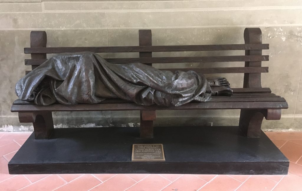 Statua in bronzo. Un corpo molto magro è avvolto in una coperta di lana grezza. Sembra rannicchiato per il freddo e per riuscire ad entrare sulla panchina. Dalla coperta escono due piedi segnati da due ferite.