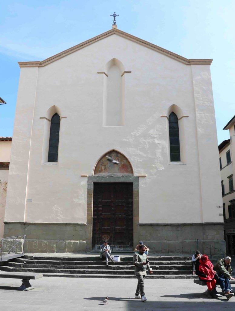 Facciata della chies di Sant'Ambrogio. Liscia senza decori se non per tre finestre. La chiesa è innalzata su di una scalinata e nella piazza davanti passeggiano alcune persone.